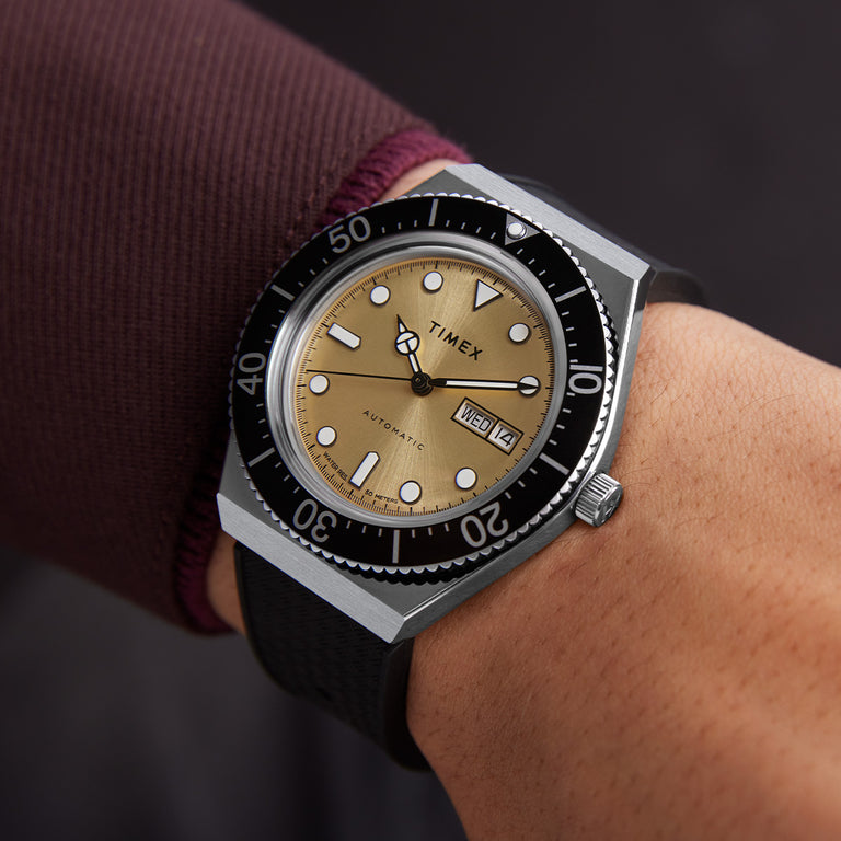 A close-up wrist shot of the Timex M79 Automatic watch, featuring a brushed champagne-gold dial with bold white hour markers and black accents. The watch has a black unidirectional bezel with white numerals and a silver stainless steel case. It displays the day and date at the 3 o'clock position. The wearer’s outfit includes a maroon long-sleeve cuff, complementing the watch’s sophisticated yet sporty aesthetic. The watch is paired with a textured black strap, emphasizing its vintage dive-inspired design.