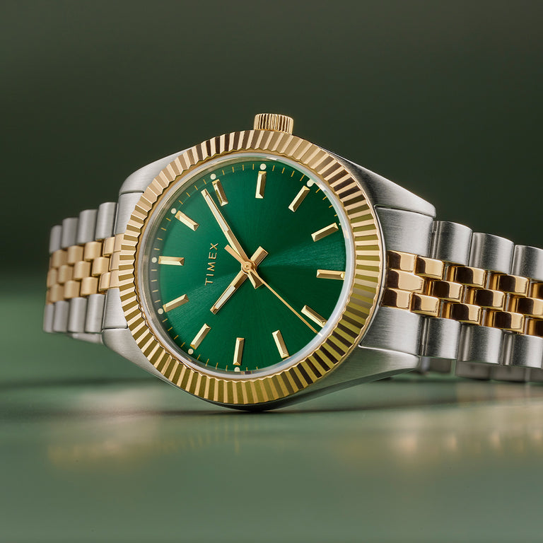 A detailed close-up of the Timex Legacy wristwatch with a striking emerald green sunray dial and polished gold-tone hour markers and hands. The watch features a fluted gold-tone bezel and a two-tone stainless steel bracelet with alternating silver and gold links. The brushed and polished finishes create a refined contrast, enhancing the watch’s classic yet modern design. The timepiece is displayed against a soft green gradient background, emphasizing its sophisticated and elegant aesthetic.