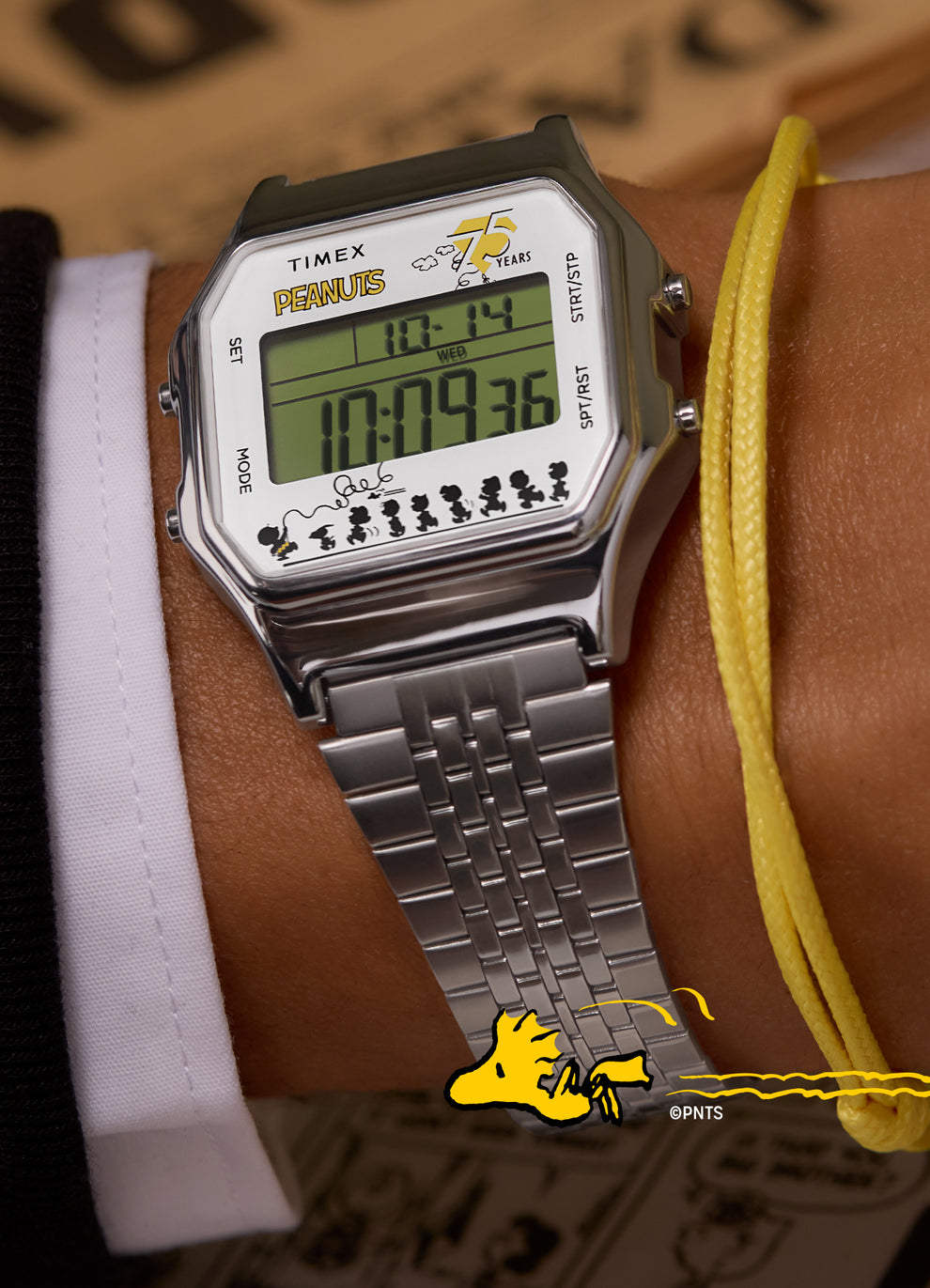 A person wearing the Timex x Peanuts® T80 watch on their wrist. The watch’s digital screen displays the time and date, while the Peanuts® branding and characters add a playful touch. A yellow cord bracelet is also visible, complementing the Peanuts® theme.